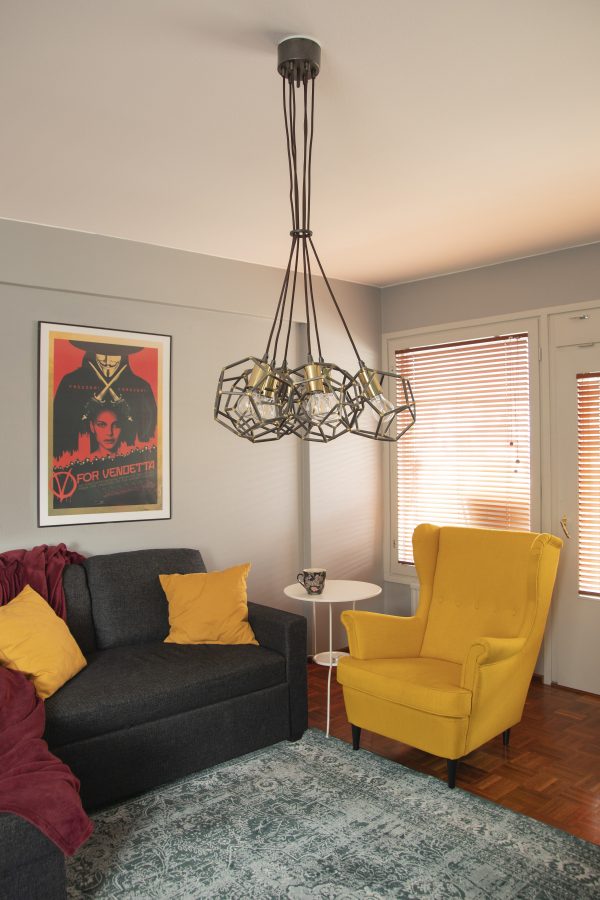 Olohuoneen sisustus jossa metallinen kuusiosainen Nora kattovalaisin, harmaa sohva, keltainen nojatuoli, taulu pöytä ja matto.
