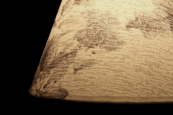 Valokuva lampunvarjostimesta, jonka sisällä palaa valo, joka tuo esille kankaan pinnassa olevan kuvioinnin. Varjostin näkyy kuvassa beigen sävyisenä. Tausta on musta. Valaisimessa on tummaa kuviointia.