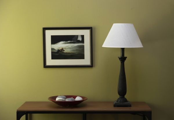 Musta puinen pöytävalaisin ja vaalea varjostin sivupöydällä. Taulu seinällä.