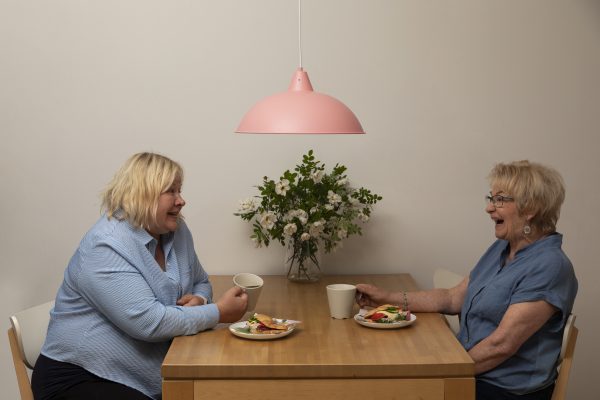 Keittiön sisustus, jossa on pinkki Lulu kattovalaisin, vaalealla pöydällä kukkia ja astioita. Kaksi naista istuu pöydän ääressä juomassa kahvia.