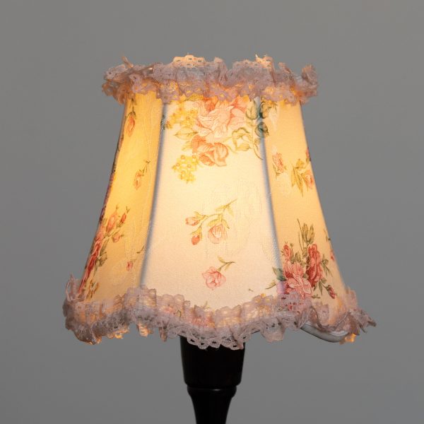Vaalea kukkakuvioinen lampunvarjostin, jonka reunoissa pitsikoriste.